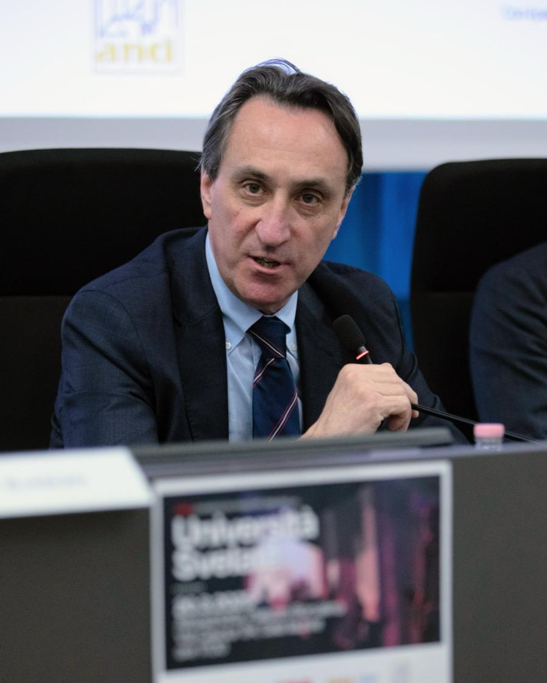 Il Magnifico Rettore dell'Università del Piemonte Orientale Menico Rizzi mentre parla al microfono seduto al tavolo.